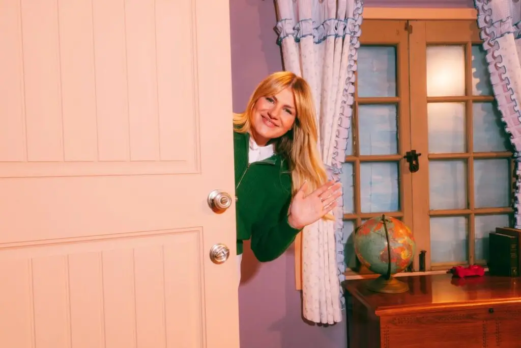Maddie Zahm peeks around a door smiling and waving in a Mister Rogers-style set