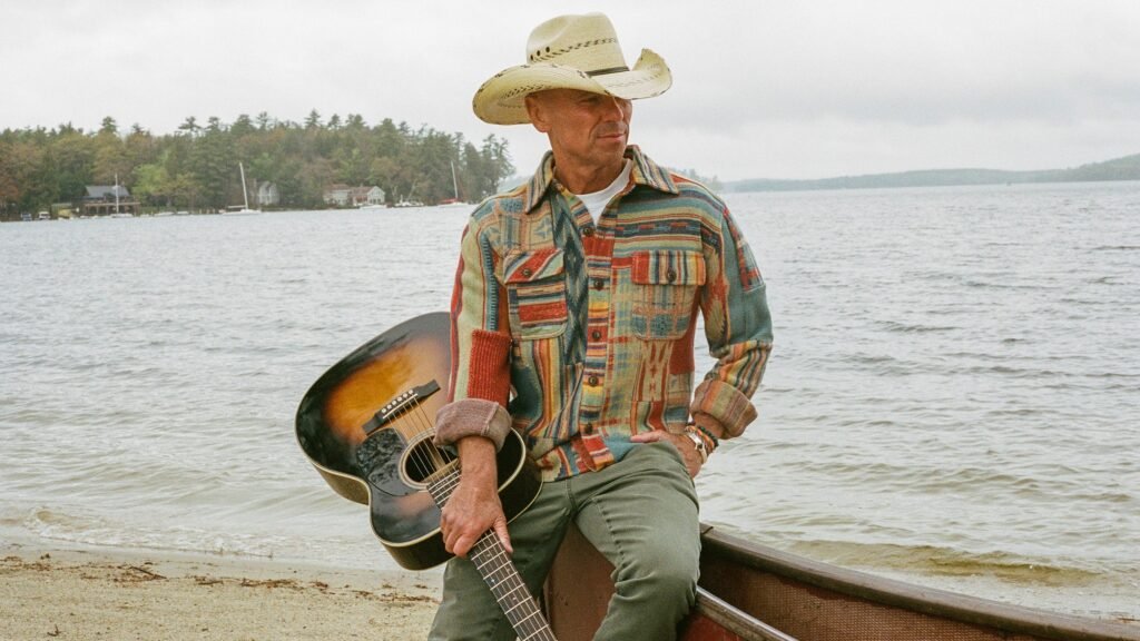 Kenny Chesney with acoustic guitar seated on a canoe at a lakeside, wearing cowboy hat
