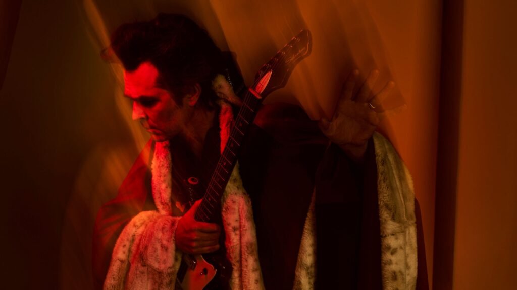 Jon Spencer playing electric guitar, wearing fur coat under dramatic red stage lighting