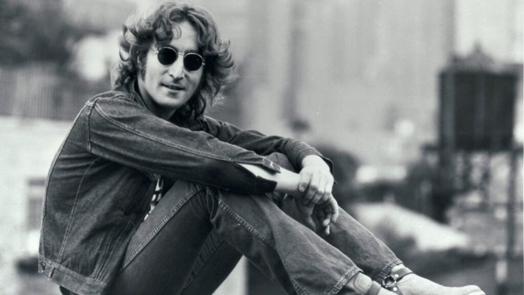 John Lennon seated on rooftop in denim jacket and round sunglasses, black and white archival photo