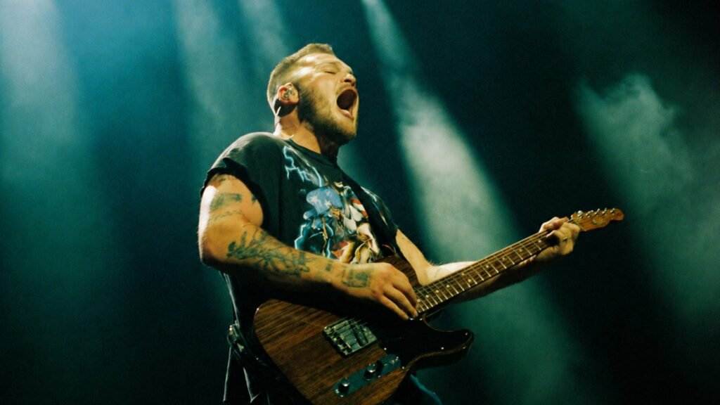 Zach Bryan performing live, mid-song with guitar, on stage with blue concert lighting