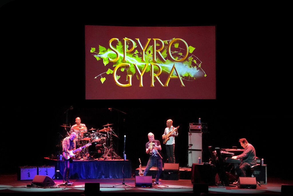 Spyro Gyra performing live on stage, saxophonist Jay Beckenstein center-front with full band behind Spyro Gyra backdrop