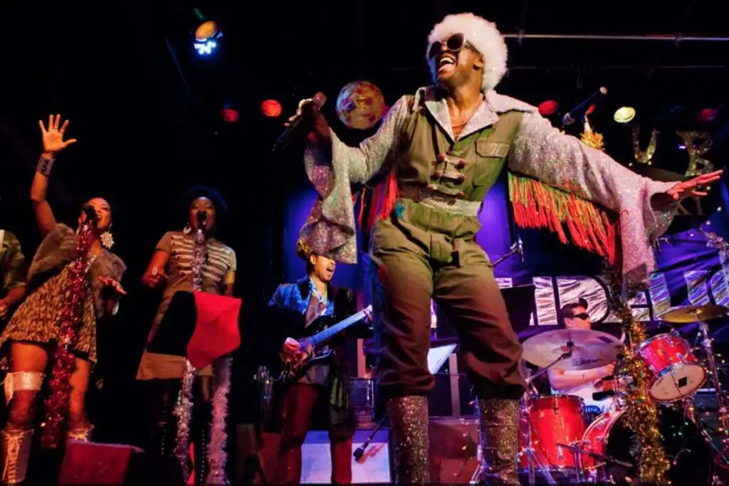 Everett Bradley as Papadelic performing with the Holidelic band, leaping on stage in glitter costume and white afro