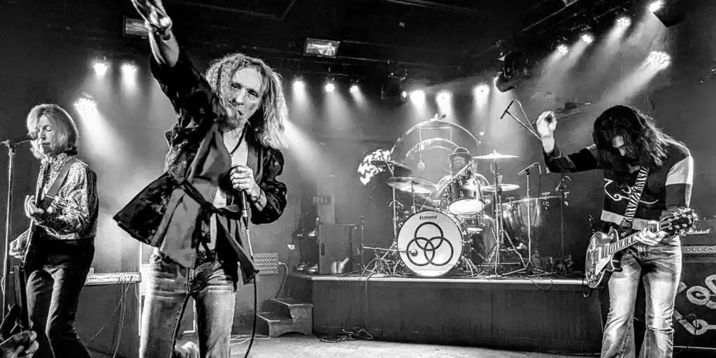 Houses of the Holy performs live on stage, vocalist raising arm mid-song with full four-piece band behind Ludwig kit