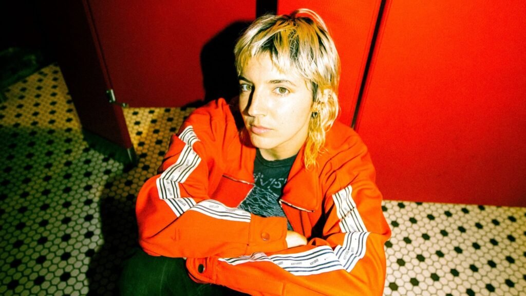 Caroline Rose official press photo, seated in red tracksuit against red walls