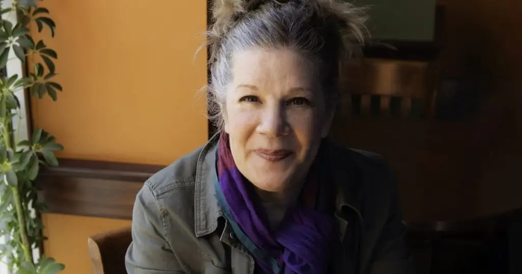 Dar Williams seated at a café table holding a coffee cup, silver hair, warm smile