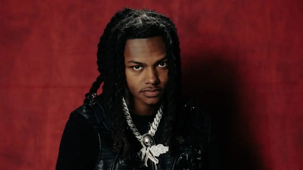 Babyfxce E in official press portrait, diamond chain and dreadlocks, deep-red studio backdrop