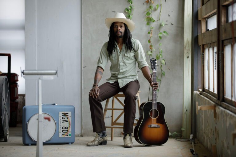 Nicky Diamonds seated with acoustic guitar in rustic industrial setting, white cowboy hat and dreadlocks