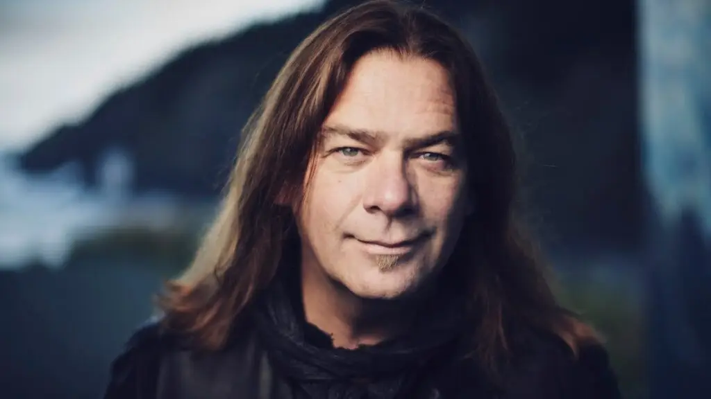 Alan Doyle smiling in a professional press photo with a rocky coastal backdrop