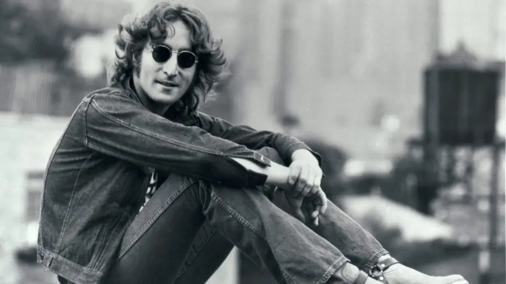 John Lennon seated in black-and-white portrait, round sunglasses and denim jacket