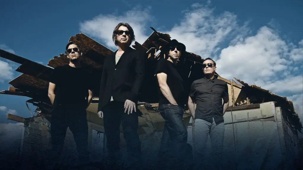 54-40 official band photo, four members posed in sunglasses against dramatic sky and industrial backdrop