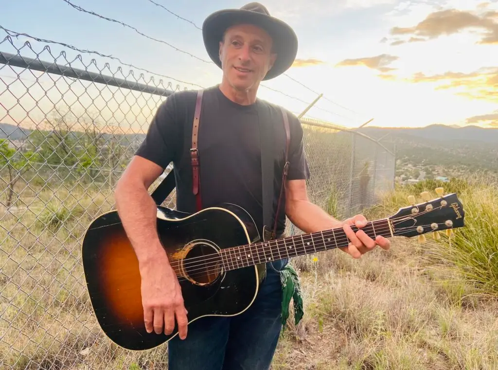 Dan Bern with acoustic guitar in outdoor press photo at dusk