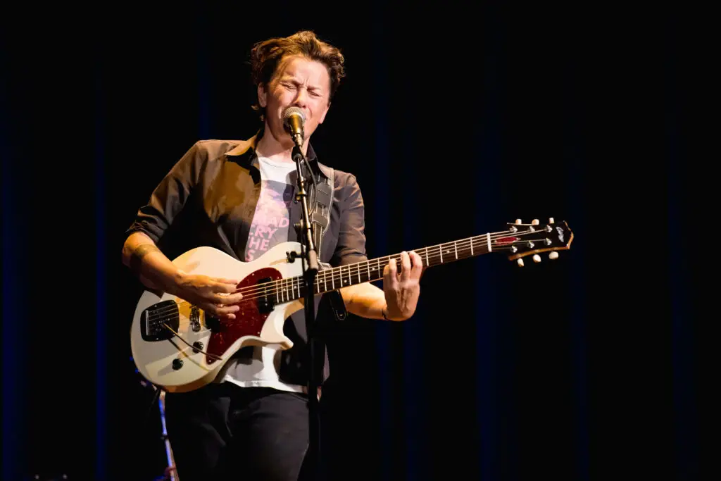 Melissa Ferrick performing live, playing electric guitar at a microphone on a dark stage