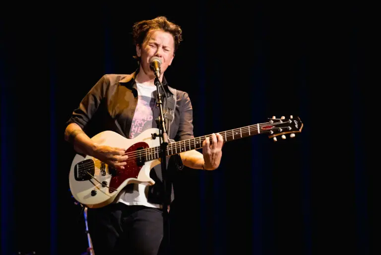 Melissa Ferrick performing live, playing electric guitar at a microphone on a dark stage
