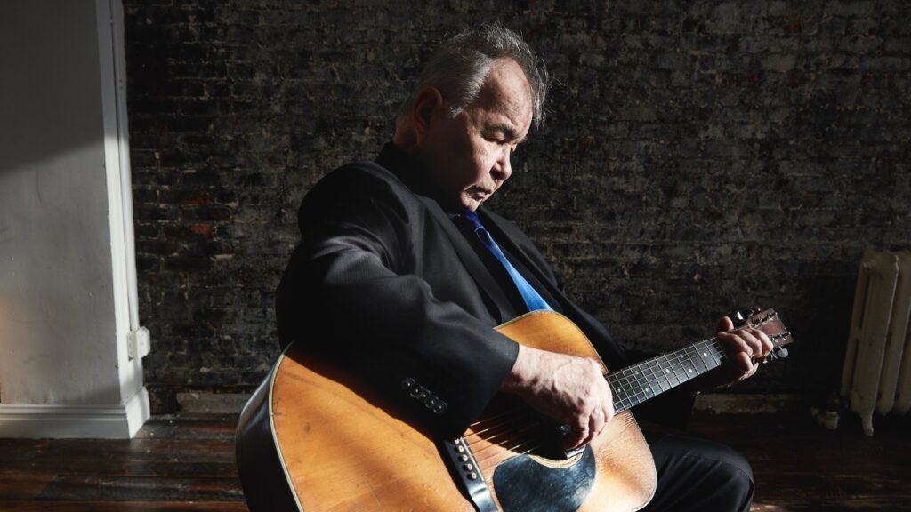 John Prine playing acoustic guitar, seated in contemplation against exposed brick wall backdrop