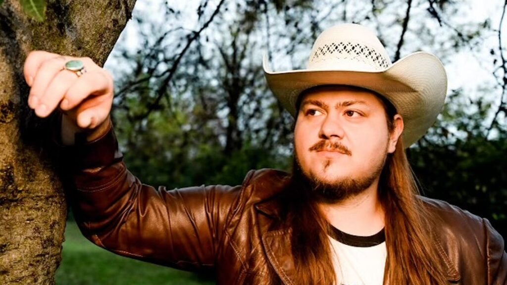 Vincent Neil Emerson in cowboy hat and leather jacket leaning against a tree, outdoor press photo